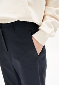 Persona con pantalones oscuros color navy y mano en el bolsillo, y camiseta de manga larga color crema con detalle en los puños, contra un fondo blanco.