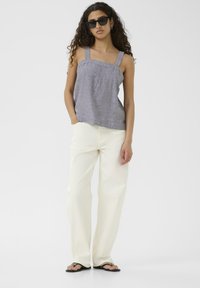 Szary top bez rękawów z kwadratowym dekoltem i plisami, połączony z białymi spodniami o szerokich nogawkach i czarnymi klapkami, na jednolitym tle.