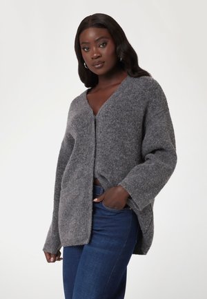 Grau, flauschige Strickjacke mit V-Ausschnitt, Knopfverschluss vorne und seitlichen Schlitzen, kombiniert mit dunkelblauen Jeans. Lässiges, lockeres Design.