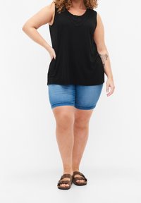 Kobieta w czarnej bezrękawnikowej bluzce, niebieskich dżinsowych szortach i brązowych sandałach, stoi z jedną ręką na biodrze na białym tle.