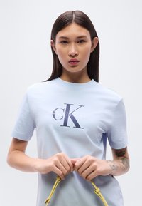 Ανοιχτό μπλε βαμβακερό t-shirt με σκούρο λογότυπο. Κοντά μανίκια και στρογγυλή λαιμόκοψη. Το μοντέλο κρατά κίτρινες κορδόνες στα χέρια.