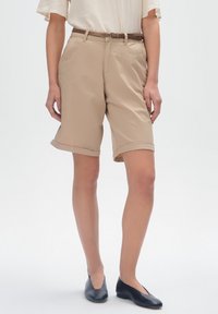Beżowe szorty do kolan z podwiniętym brzegiem, wykonane z lekkiego materiału, z przednią kieszenią i brązowym skórzanym paskiem. Płaskie czarne buty.