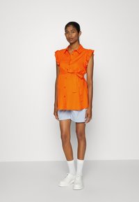 Camisa laranja de botão com mangas fruncidas, atada à cintura, combinada com calções azul claro e sapatos e meias brancas. Design limpo e moderno.