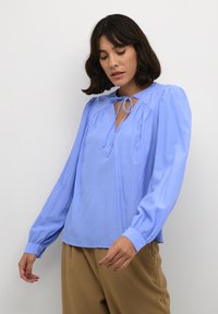 Blusa azzurra chiara realizzata in tessuto morbido, con maniche lunghe a sbuffo, scollatura a chiave con fiocco e sottili righe verticali.