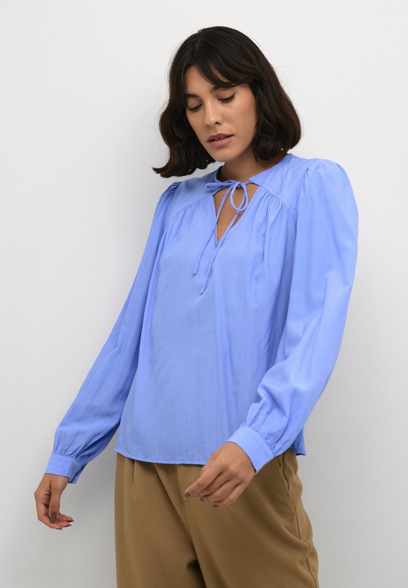 Blusa azzurra chiara realizzata in tessuto morbido, con maniche lunghe a sbuffo, scollatura a chiave con fiocco e sottili righe verticali.