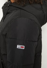 Sort puffersjakke med quiltet design, fast hætte og et lille broderet logo med rød, hvid og blå detaljering på ærmet.