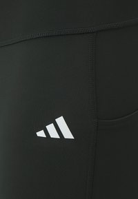 Leggings de sport noirs en matériau extensible, avec un logo à trois rayures blanches en bas à droite et des détails de coutures à plat.