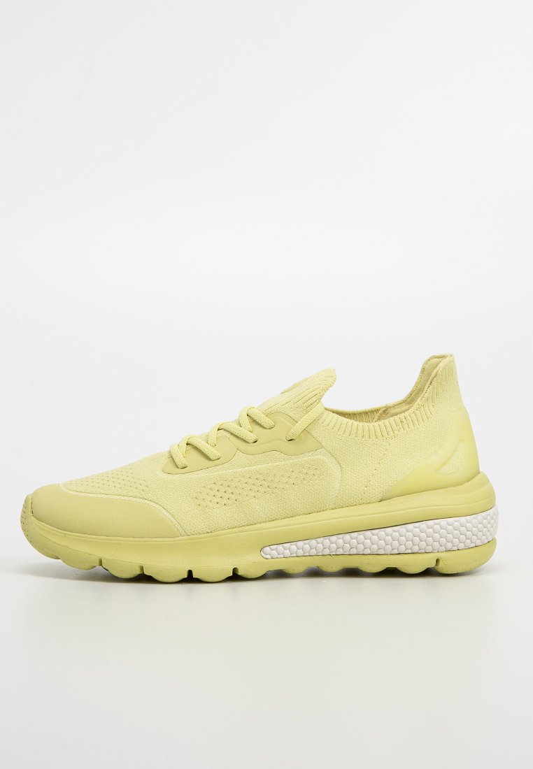 Zapatilla deportiva de color amarillo claro con una parte superior texturada, punta redonda y una suela gruesa que presenta una banda de acento blanca con puntos.