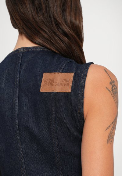 Σκούρο τζιν χωρίς μανίκια ένδυμα με καφέ ετικέτα "Baum und Pferdgarten" στο επάνω μέρος της πλάτης, τατουάριστο χέρι και μακριά καστανά μαλλιά που φαίνονται από πίσω.