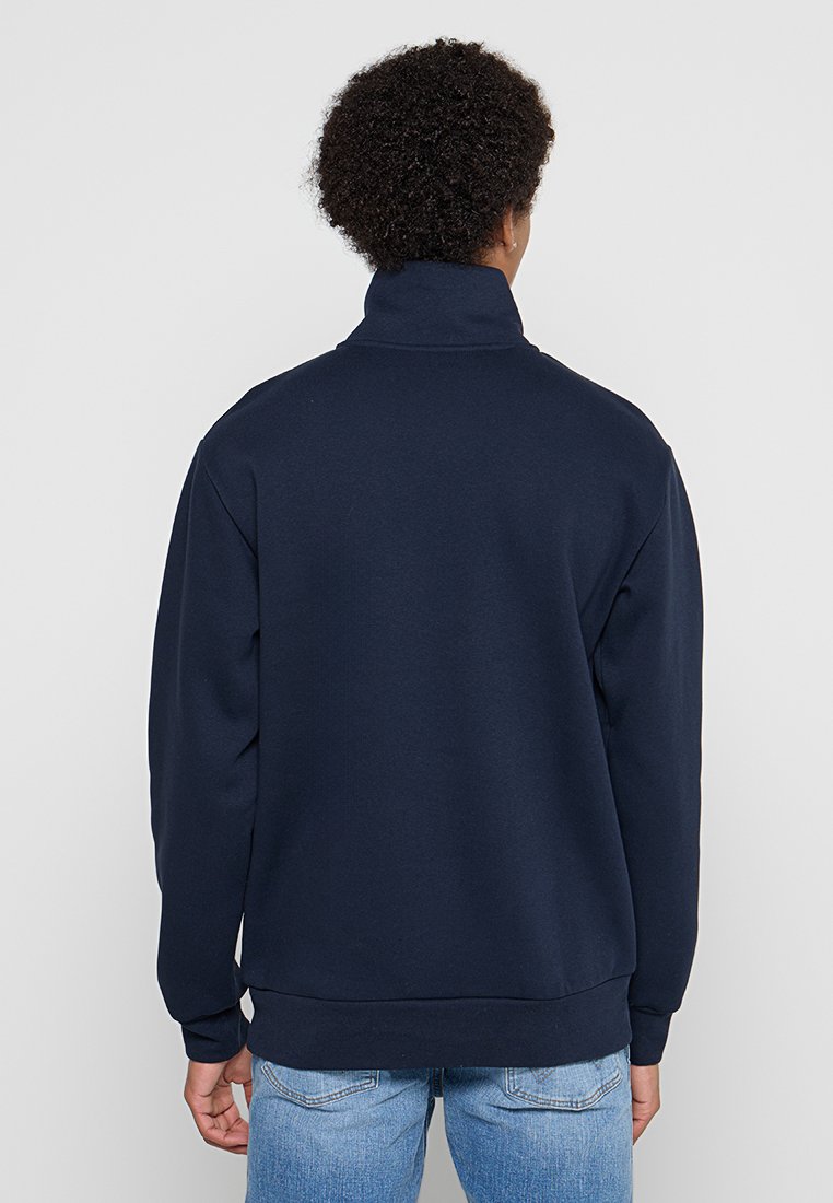 Sweatshirt bleu marine avec un col montant, des manches longues et des poignets côtelés. Présente une coupe décontractée et une texture douce, associé à un jean bleu clair.