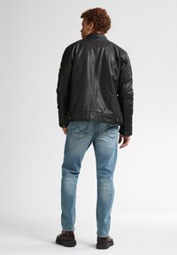 Chaqueta de cuero negra con costuras detalladas, puños con cremallera y parches, combinada con jeans azul claros y botas marrones.