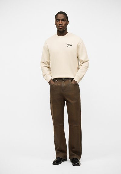 Άνδρας στέκεται με τα χέρια στις τσέπες, φοράει κρεμ φούτερ με μικρό μαύρο κείμενο και χαλαρό καφέ παντελόνι με μαύρα παπούτσια, σε λευκό φόντο.