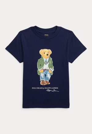 Marineblå t-shirt med en bjørn iført en grøn jakke, hvid sweater, blå jeans og brune sko, med teksten "Polo Bear by Ralph Lauren" nedenunder.