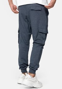 Mężczyzna w ciemnoniebieskich spodniach dresowych z kieszeniami cargo i białych sneakersach, stojący na tle jednolitego białego tła.