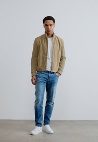 Μπεζ σατινέ μπουφάν πάνω από λευκό t-shirt, συνδυασμένο με ανοιχτό μπλε τζιν και λευκά αθλητικά παπούτσια· στέκεται μπροστά από απλό λευκό φόντο.