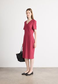 Tætsiddende midi-kjole med korte ærmer i dyb pink med V-hals, lavet af struktureret stof. Sammen med sorte flade sko og en sort håndtaske.