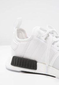 Vit nätliknande sportskor med ribbad svart sula, vadderad krage och vita snören. Har tre vita ränder på sidan och en dragflik.