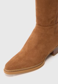 Bottines en suède marron à bout pointu, avec des détails de couture subtils et un petit talon en bois. Texture lisse avec une finition mate.