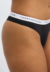 Μαύρο εσώρουχο στρινγκ με λευκή ελαστική ταινία μέσης που φέρει το λογότυπο "TOMMY HILFIGER". Μαλακό ύφασμα με ομαλή υφή.