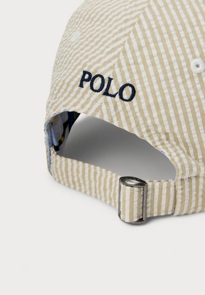 Șapcă bej cu dungi verticale, brodat cu „POLO” în albastru închis. Dispune de cataramă metalică reglabilă și material texturat.