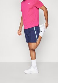 Herrarnas sportoutfit med en ljusrosa T-shirt med kort ärm och marinblå shorts med en ljusblå sidostrand, tillsammans med vita sneakers och strumpor.