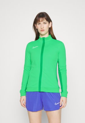 Groene ritssluitjak gemaakt van rekbare stof, voorzien van een hoge kraag en een klein Nike-logo op de linkerkant van de borst. Gecombineerd met paarse shorts.