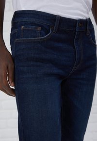Jean en denim bleu foncé avec un design classique à cinq poches, un léger effet vieilli et des coutures renforcées le long des coutures.