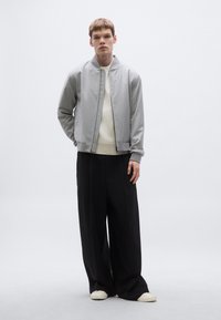 Szürke bomber dzseki könnyű anyagból, bordázott mandzsettákkal és cipzáras lezárással, fehér bordázott pulóverrel és fekete, bőszárú nadrággal párosítva.