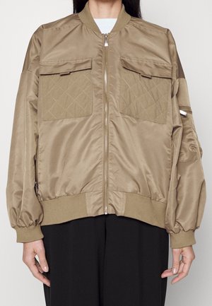 Chaqueta bomber de satén marrón claro con bolsillos con solapa acolchados, cremallera delantera, puños y dobladillo acanalados, usada sobre una camiseta blanca y pantalones negros.