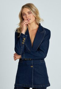 Blazer in denim blu scuro, con un design aderente, rever a V e sei bottoni dorati sulla manica sinistra come dettagli decorativi. Tessuto liscio.