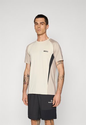 Jeune homme aux cheveux courts et foncés, portant un t-shirt de sport beige et noir de BOSS et un short noir de Sergio Tacchini, se tenant contre un fond clair uni.