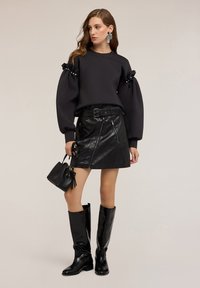 Donna in top nero con maniche a sbuffo e dettagli di perle, gonna in pelle nera con cintura, stivali neri fino al ginocchio, grandi orecchini d'argento e che tiene una borsa nera.