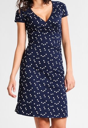 Kobieta ubrana w granatową sukienkę z krótkim rękawem, sięgającą kolan, ozdobioną białymi wzorami stokrotek i z dekoltem w kształcie litery V, owijaną.
