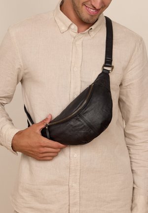 Schwarze Leder-Gürteltasche mit geschwungener Form, mit Reißverschluss versehenem Frontfach und verstellbarem Riemen, die über einem hellen Hemd quer über den Körper getragen wird.