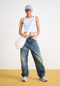 Borsa a spalla bianca testurizzata con una tracolla attorcigliata, top senza maniche azzurro chiaro e jeans blu larghi, in piedi su uno sfondo giallo.