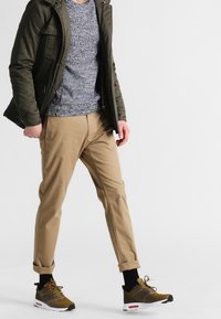 Oliwkowa zielona kurtka, teksturowany niebieski sweter, zwężane spodnie khaki oraz brązowe tenisówki z czerwonymi akcentami. Casualowy strój z podwiniętymi mankietami.