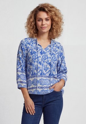 Vrouw met krullend blond haar die een blauw-wit blouse met patroon en driekwart mouwen draagt en donkerblauwe spijkerbroek, hand in zak.