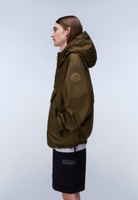 Ung person i profil iført en olivengrøn hættetrøje med logo-patch på ærmet og sorte cargoshorts med stor lomme.