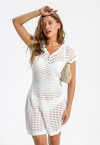 Robe en maille blanche présentant un design en maille ajourée, manches courtes et taille cintrée. Détails avec des boutons et un motif de tissu texturé.