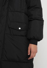 Manteau noir rembourré avec une texture matelassée, une poche latérale avec un bouton-pression argenté et des poignets côtelés.