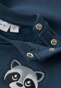 Felpa blu navy con collo a coste, bottoni a pressione argentati e ricamo di un procione grigio sulla parte frontale. Tessuto interno morbido.