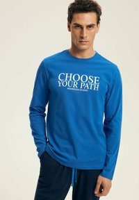 Man wearing a blue long-sleeve shirt with "CHOOSE YOUR PATH HENDERSON STUDIOS" text, paired with navy pants, standing against plain background.