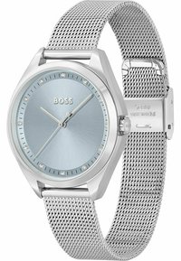 Reloj de malla plateada con una esfera sol de color azul claro. Presenta marcadores de diamante, manecillas de horas y minutos en negrita, y un bisel pulido. Logotipo de la marca exhibido.