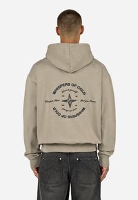 Felpa beige con cappuccio e cordino, caratterizzata da una stampa grafica sul retro che include una bussola e testo in nero. Texture del tessuto morbida.