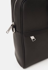 Un sac à dos en textured synthétique noir avec des détails de fermeture éclair argentés et une étiquette de marque. La forme est rectangulaire avec une base légèrement arrondie.