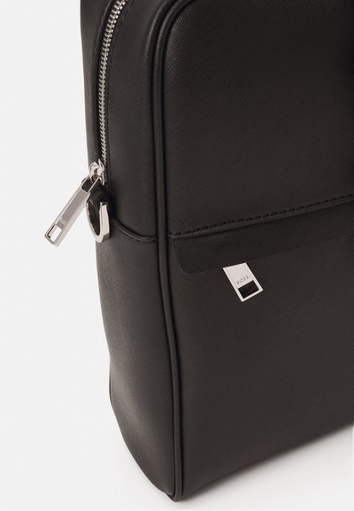 Un sac à dos en textured synthétique noir avec des détails de fermeture éclair argentés et une étiquette de marque. La forme est rectangulaire avec une base légèrement arrondie.