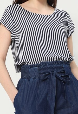 Femme portant un haut à manches courtes rayé vertical noir et blanc rentré dans un pantalon taille haute bleu marine style paperbag avec une ceinture nouée.