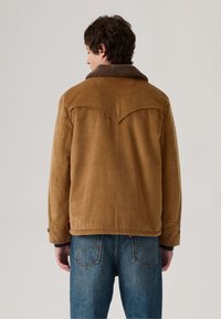 Chaqueta de pana marrón con un cuello de borreguito, costuras visibles, puños abotonados y un diseño de yugo en la parte posterior. Combinada con jeans de mezclilla azules.