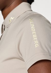Beige polo shirt med korte ærmer og struktureret stof. Har et gult logo på venstre skulder og en krave.