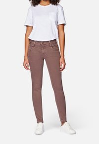 Mujer vestida con una camiseta blanca con bolsillos, jeans ajustados color malva y zapatillas blancas, de pie con las manos relajadas frente a un fondo blanco.